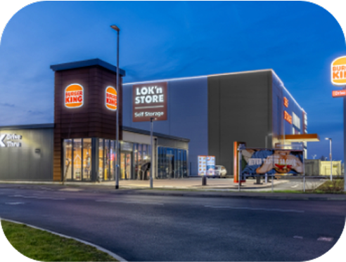 A Burger King restaurant and a Lokn Store self-storage facility are side by side, illuminated at dusk with clear signage and a mostly empty parking lot in front.