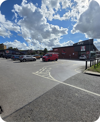 A KFC restaurant with a drive-thru is shown under a partly cloudy sky. Several cars and a red van are parked in the large parking lot. The area is well-lit by sunlight.