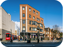 A red brick, four-story corner building with shops on the ground floor sits on a busy street with pedestrians and a red double-decker bus, under a clear blue sky.