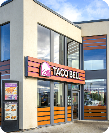 The exterior of a modern Taco Bell restaurant with large windows, an illuminated Taco Bell sign, and a drive-thru menu board visible on the left side of the building.