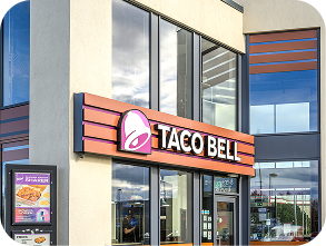 Taco Bell restaurant exterior with a large logo sign above the entrance and a digital menu board displaying food items on the left side of the building.