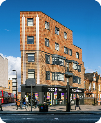 A modern brick building on a street corner features a Taco Bell restaurant on the ground floor. People are walking by, and a red double-decker bus is visible on the left. The sky is blue with some clouds.