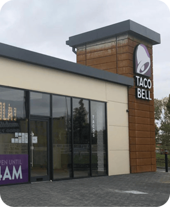 A Taco Bell restaurant with a tall sign displaying the Taco Bell logo and name. The building has large glass windows and a banner indicating it is open until 4 a.m. Trees and a cloudy sky are visible in the background.