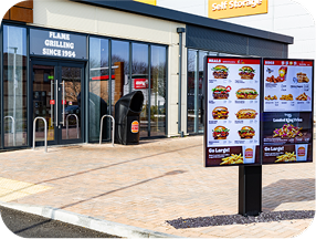 A fast food restaurant with glass doors is shown, featuring a drive-thru menu displaying images of burgers, meals, and drinks. A sign above the entrance reads FLAME GRILLING SINCE 1954.
