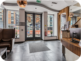 Modern hotel lobby with large glass doors, a curved reception desk with computer monitors, brown seating area, and large windows showing a city street with buildings and pedestrians outside.