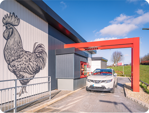A white car is stopped at a drive-thru window of a modern restaurant with a large black-and-white rooster mural on the exterior wall, under a bright blue sky.